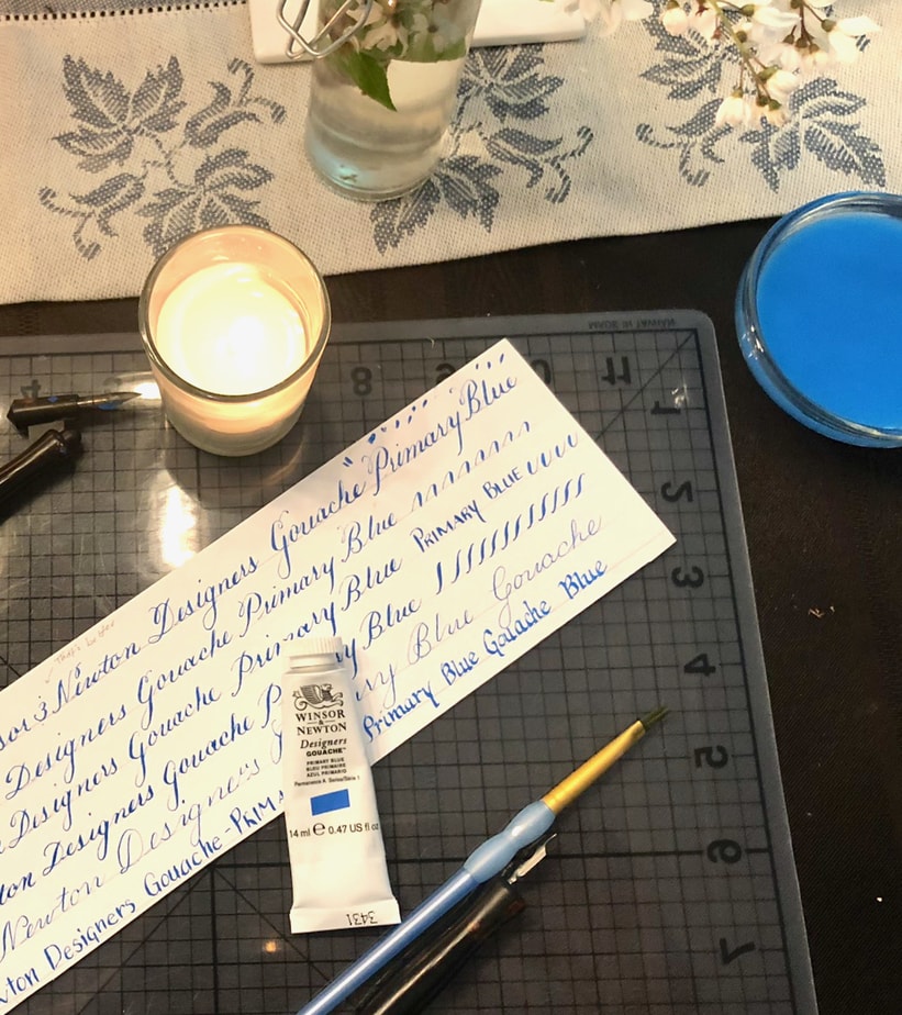 Hand-drawn Copperplate, Spencerian, and block calligraphy on white paper using Winsor & Newton Designers Gouache in Primary Blue. A Speedball oblique pen at the top of the page next to a decorative candle, a tube of the gouache is shown in the center of the page, and a paintbrush and straight pen are shown at the bottom of the image. The items sit on a self-healing mat.