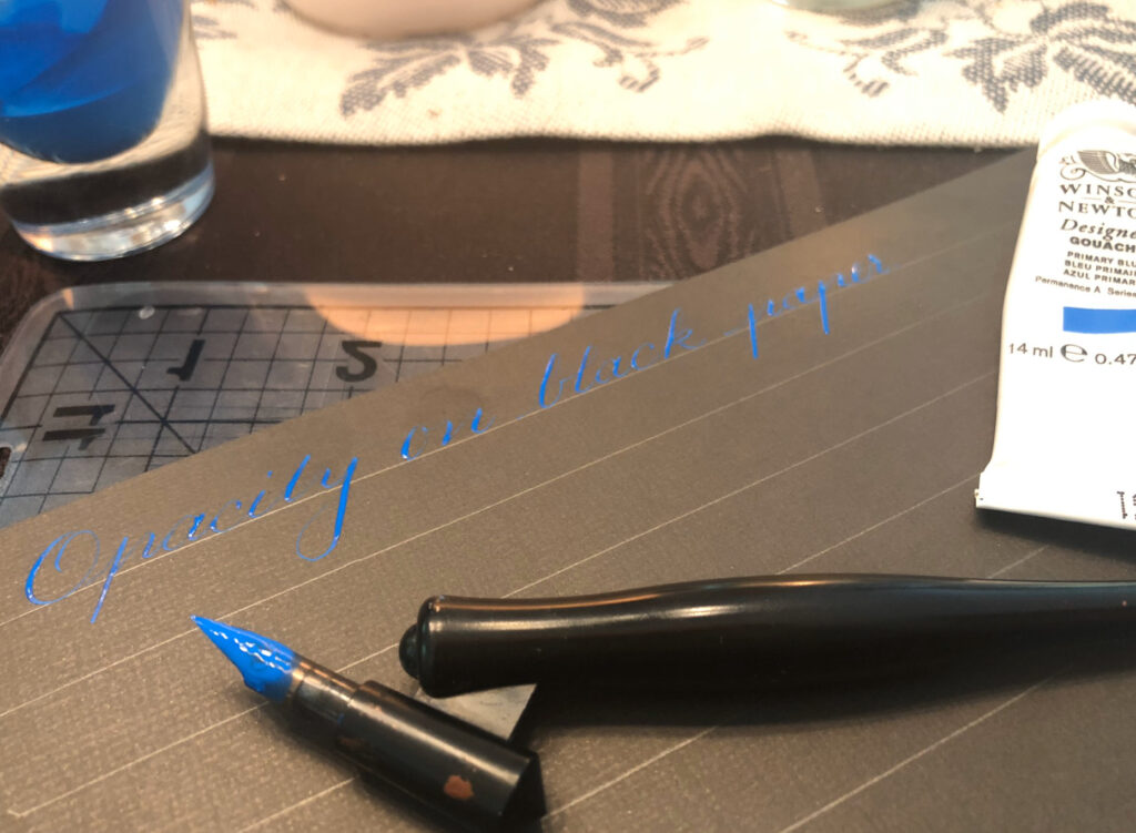 Hand-drawn Spencerian calligraphy on black paper using Winsor & Newton Designers Gouache in Primary Blue to show the product’s high opacity. A Speedball oblique pen at the bottom of the page next t a tube of the gouache.